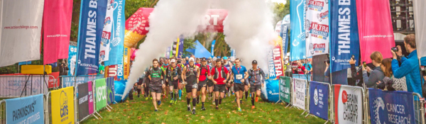 People are walking between events barriers. The barriers include banners and flags with different charity and organisation branding. A fog machine is being used in the distance. 