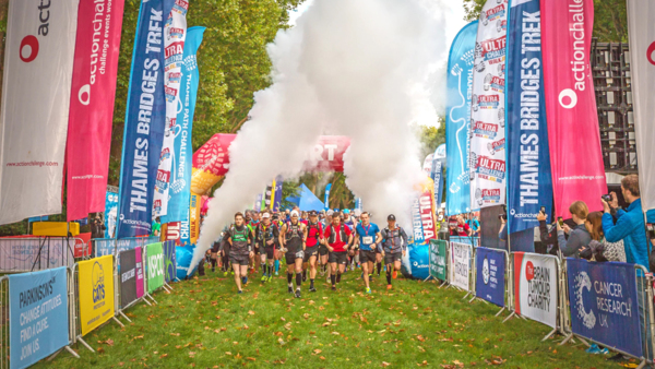 People are walking between events barriers. The barriers include banners and flags with different charity and organisation branding. A fog machine is being used in the distance. 
