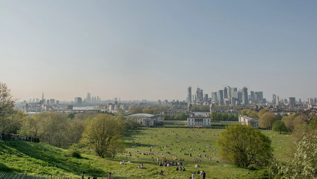 A landscape view of Greenwich Park in London. The image was taken on top of a hill at the park. People are scattered across the park and Queen's House can be seen. In the distance there is the skyline of central London.