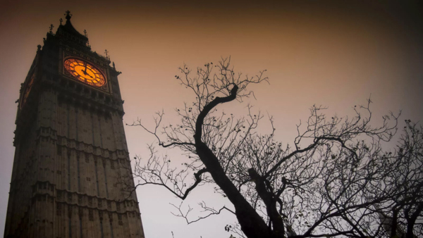 Pictured is Big Ben, the famous clock tower in London. The clock face is tinted orange. In front of the tower is a tree with no leaves. The image is dark and likely taken in the evening.