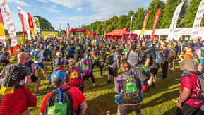 A crowd of people are standing near an event starting line. There are event flags on either side and in the distance a start line. There are event card numbers on people's backpacks and backs.