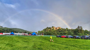 A person in a yellow jacket and black shorts is walking across a field. On the right there are a row of parked cars. On the left is a festival set up with tents and flags. In the background there is a castle and a rainbow. 