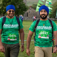 Two men are walking. They are wearing green Macmillan tops with an event number pinned to the tops. They are also wearing dark blue pagris and backpacks. 