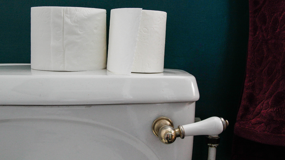 View of a toilet that shows the toilet handle and top of the toilet. On top is two rolls of toilet paper, one has been used. The wall behind is a dark blue.