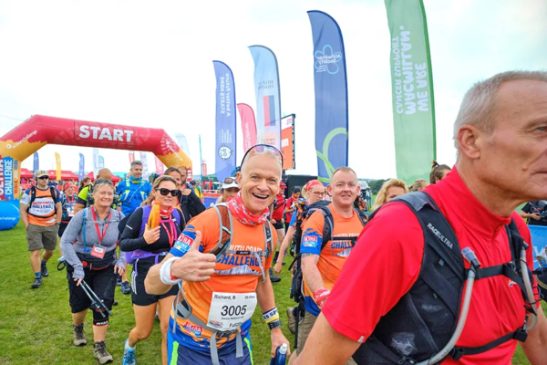 A man in a bright orange top is smiling and holding a thumb's up. He is in a crowd of other people who are walking. Behind him is a balloon arch with the words start on it. 