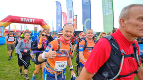 A man in a bright orange top is smiling and holding a thumb's up. He is in a crowd of other people who are walking. Behind him is a balloon arch with the words start on it. 