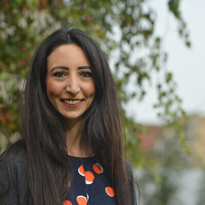 Dessie has long black hair and is smiling. She is wearing a orange polka dotted top and a dark grey cardigan. The background behind her is blurred. Dessie is a Macmillan Cancer Voice.
