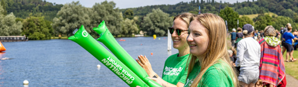 Two people wearing green Macmillan t-shirts are standing on grass near a river. One person is holding inflatable Macmillan signs in both hands. The person behind them has their hair pulled back and is wearing black sunglasses. 