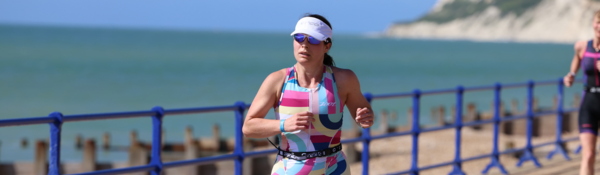 A woman wearing a brightly coloured bodysuit is running. She is also wearing a white sun visor and sunglasses. She appears to be running on a path that is alongside a beach.