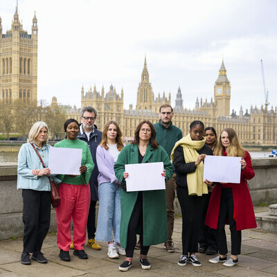 A group of people standing on a bridge near the UK Parliament building in London. The group is diverse and includes people from different age groups and backgrounds. Some people in the photo are holding white pieces of paper. 