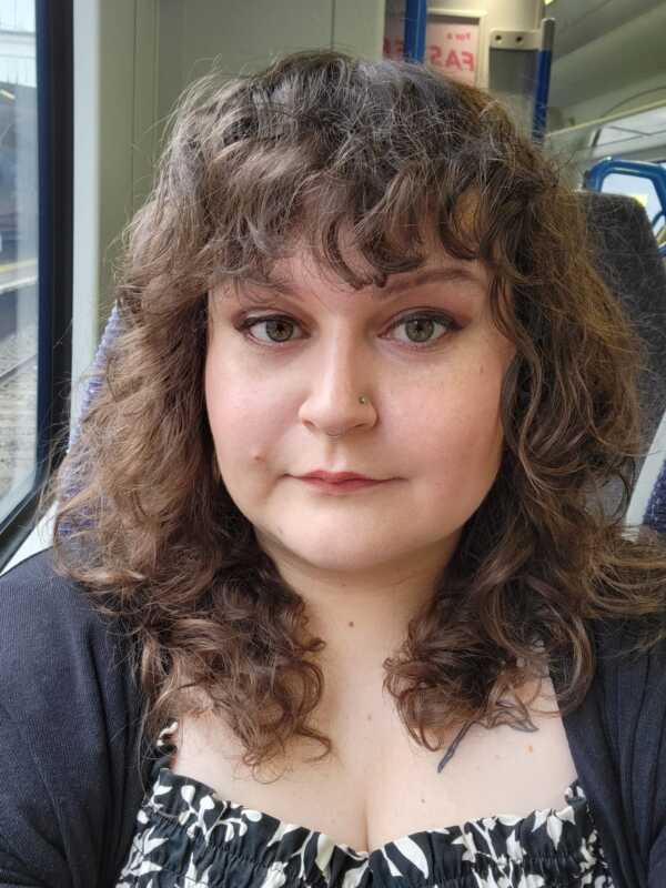 A selfie of Kerstin. Kerstin has short, brown curly hair and brown eyes. She is wearing a black cardigan and a black and white top. She is looking at the camera and appears to be sitting on a train.