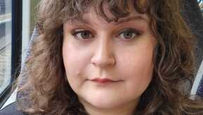 A selfie of Kerstin. Kerstin has short, brown curly hair and brown eyes. She is wearing a black cardigan and a black and white top. She is looking at the camera and appears to be sitting on a train.