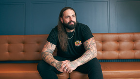 A man with long dark hair, a beard and arm tattoos sitting on the brown leather couch.