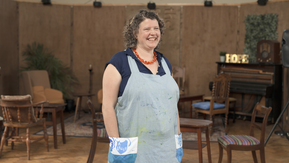 A woman standing in a room with her hands in her apron pockets covered in paint.