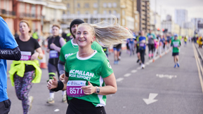 A woman is running in a green Macmillan top. Her blonde hair is pulled back in a pony tail. She appears to be running on a road, and there are other runners next to and behind her.