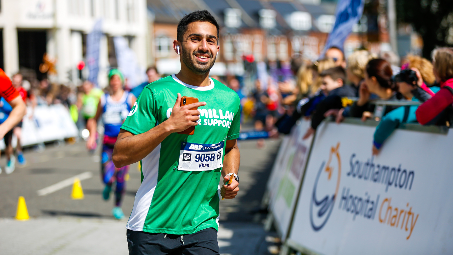 A man with short black hair is running on a road. He appears to be at an event and is wearing a green Macmillan top with an event number pinned to it. Next to him are people watching and cheering on runners. Behind him are more runners. 