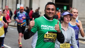 A man in a green Macmillan top is running. He has both his hands up and is giving a thumb's up. He has short dark hair. Next to and behind him are other runners. 