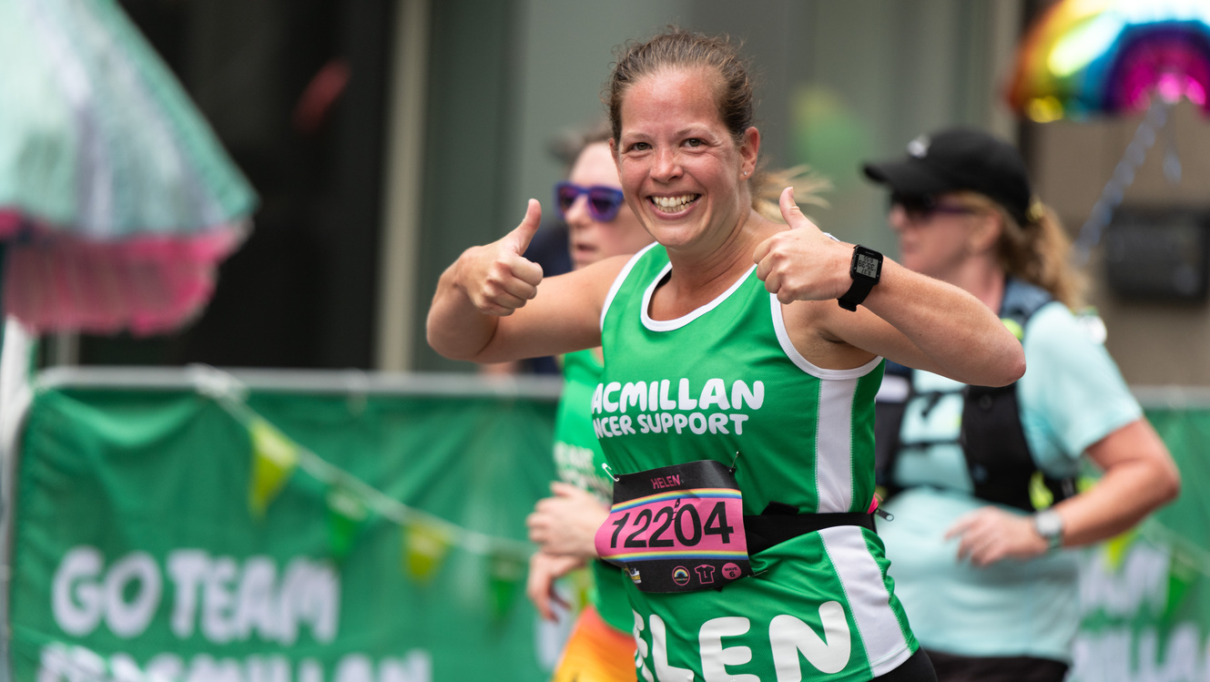 A woman is giving two thumb's up. Her brown hair is pulled back into a pony tail. She is wearing a green Macmillan top and appears to be participating in a running event. 