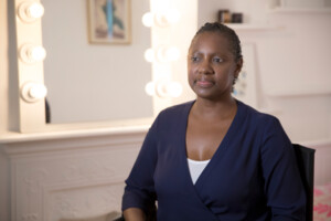 Woman in blue top sitting on chair, behind her is a mirror with lightbulbs around the mirror frame.