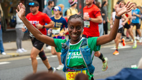 A woman running with her arms in the air. Her hair is platted and she is wearing a green Macmillan top. Behind her are other runners. 