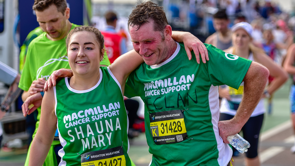 Two people wearing green Macmillan tops are standing beside one another. They each have an arm extended to wrap around the other. The person on the left has their brown hair pulled back in braids. The person on the right has short brown hair. 