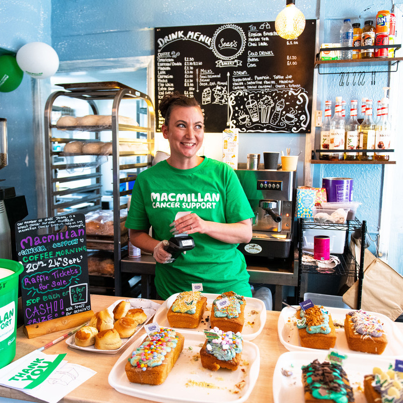 Woman in cafe wearing green Macmillan tshirt in front of her is a range of coffee morning cakes.