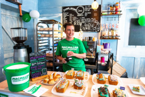 Woman in cafe wearing green Macmillan tshirt in front of her is a range of coffee morning cakes.