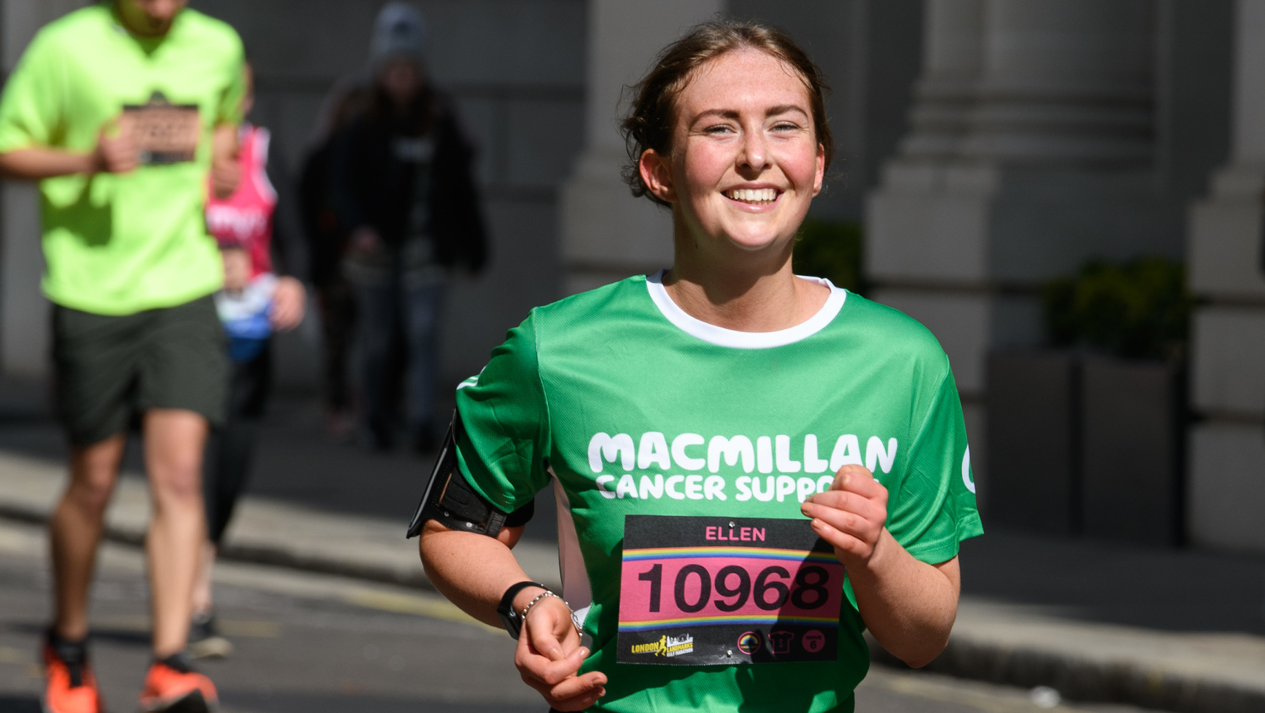 A woman is running on a road. She is wearing a green Macmillan running top and short, black running shorts. Her brown hair is pulled back and she is smiling. Pinned to her running top is a race number.
