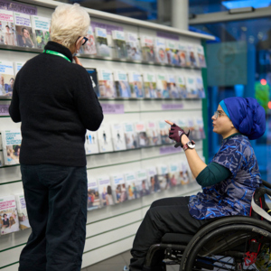Woman in wheelchair and woman in black clothing looking at cancer booklets displayed on a wall.