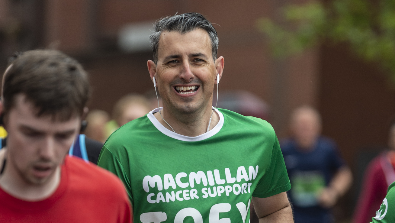 A man running at an event. He is wearing a green Macmillan running top and black shorts. He is smiling and has headphones in. Around him are other runners.