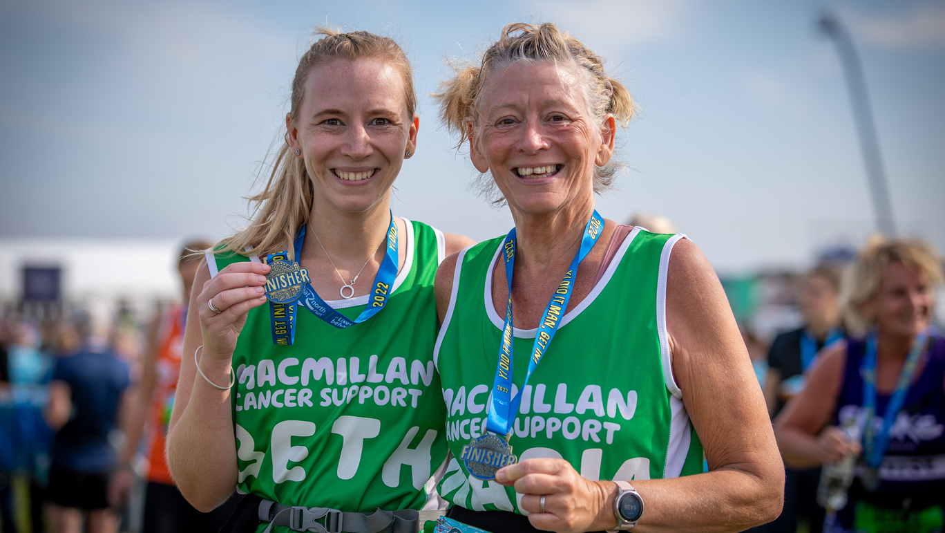 Two people smiling at a camera after finishing an event. Both are wearing green Macmillan tops and event metals around their necks. Behind them are other event participants.