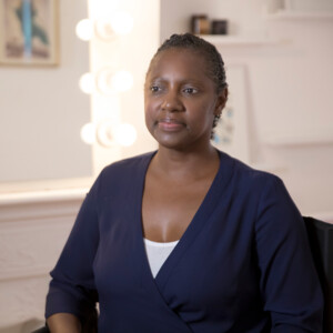 Woman in blue top sitting on chair, behind her is a mirror with lightbulbs around the mirror frame.