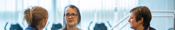 Two nurses in dark blue uniforms are sitting down. The nurses are from the Macmillan Prehabilitation Team. They are facing towards a patient who is also sitting down. The patient has on a long grey top and grey dungarees. Behind them are exercise machines. 