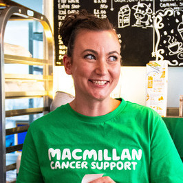 Woman in cafe wearing green Macmillan tshirt in front of her is a range of coffee morning cakes.
