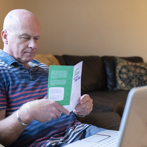 Steven sits on a sofa reading a cancer information leaflet. A laptop is open on his lap.