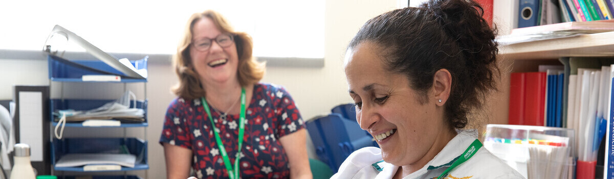 Two Macmillan professionals are sitting in an office and appear to be laughing. One person is in a white outfit and appears to be reading a document. Behind them another person in a colourful top is sitting at a desk laughing.