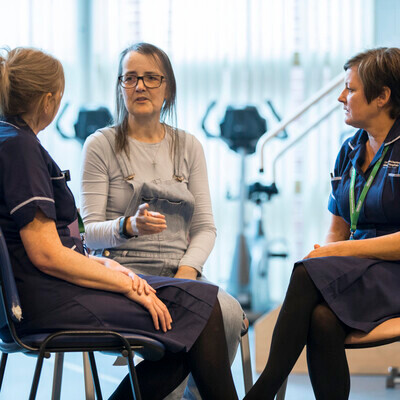 Two nurses in dark blue uniforms are sitting down. The nurses are from the Macmillan Prehabilitation Team. They are facing towards a patient who is also sitting down. The patient has on a long grey top and grey dungarees. Behind them are exercise machines. 