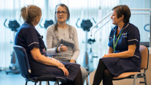 Two nurses in dark blue uniforms are sitting down. The nurses are from the Macmillan Prehabilitation Team. They are facing towards a patient who is also sitting down. The patient has on a long grey top and grey dungarees. Behind them are exercise machines. 