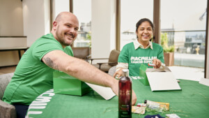 Two people are sitting at a table and packing paper boxes. Both individuals are wearing a green top. The person on the left is reaching their arm across the table to grab something and is smiling. The person on the right is putting something into a box and is smiling.