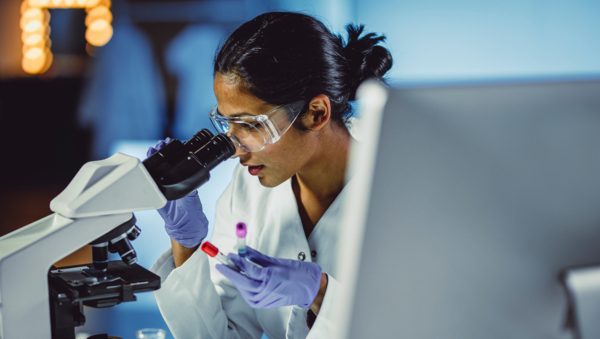 A woman in a lab looking into a microscope, holding two small blood test tubes in her left hand. 