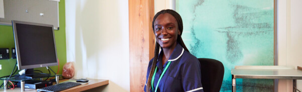 A nurse is sat in an office chair and facing towards the camera smiling. She has long braids in her hair, is wearing a blue nurse's uniform, and has on a green lanyard. Next to her is a desk with a computer. 