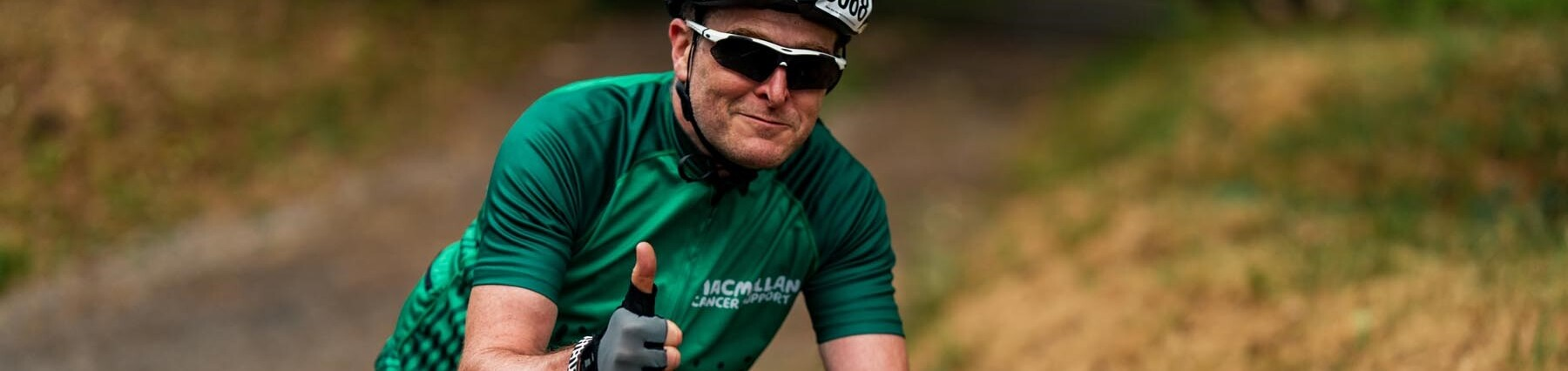 Man riding his bike with a Macmillan cycling jersey on and giving a thumbs up to the camera
