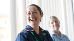 A nurse is smiling. Her brown hair is pulled back in a bun and she is wearing a dark blue NHS uniform. Behind her is another healthcare professional who is wearing a light blue NHS uniform. They also have their blonde hair pulled back. 
