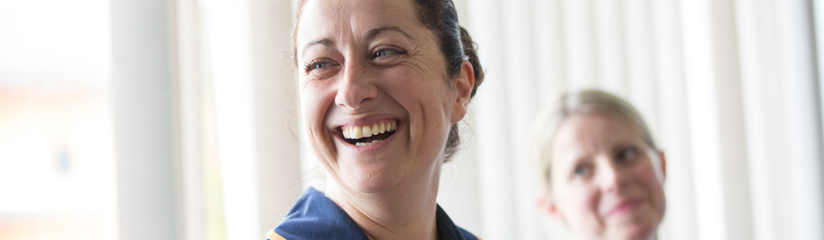 A nurse is smiling. Her brown hair is pulled back in a bun and she is wearing a dark blue NHS uniform. Behind her is another healthcare professional who is wearing a light blue NHS uniform. They also have their blonde hair pulled back. 
