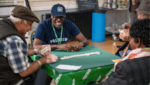 A group of people are sitting around a table playing dominoes. The tap has a green Macmillan logo table top on it. 
