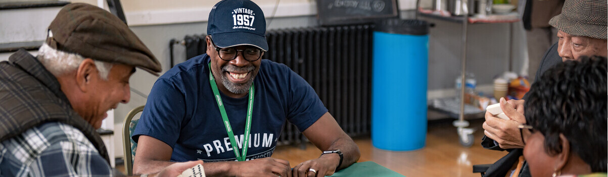 A group of people are sitting around a table playing dominoes. The tap has a green Macmillan logo table top on it. 