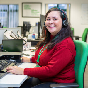 Jenny is a Macmillan Support Line Welfare Rights Advisor. She is sitting at a desk and smiling at the camera. She is wearing a red top and blue jeans. On her head is a headset and she appears to be working at a computer. 