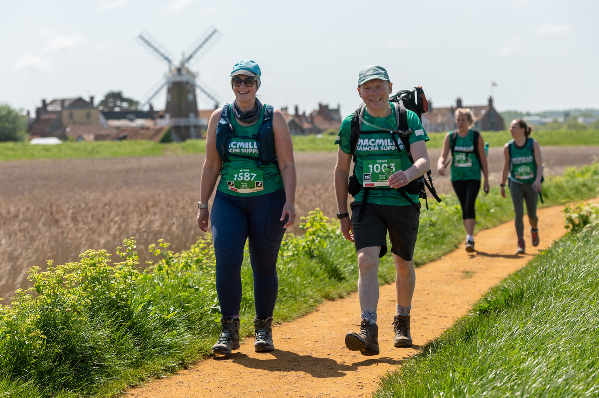 Participants are walking the Norfolk Coast Mighty Hike. Both are wearing green Macmillan tops and caps. Behind is a view of a local village and other participants. 