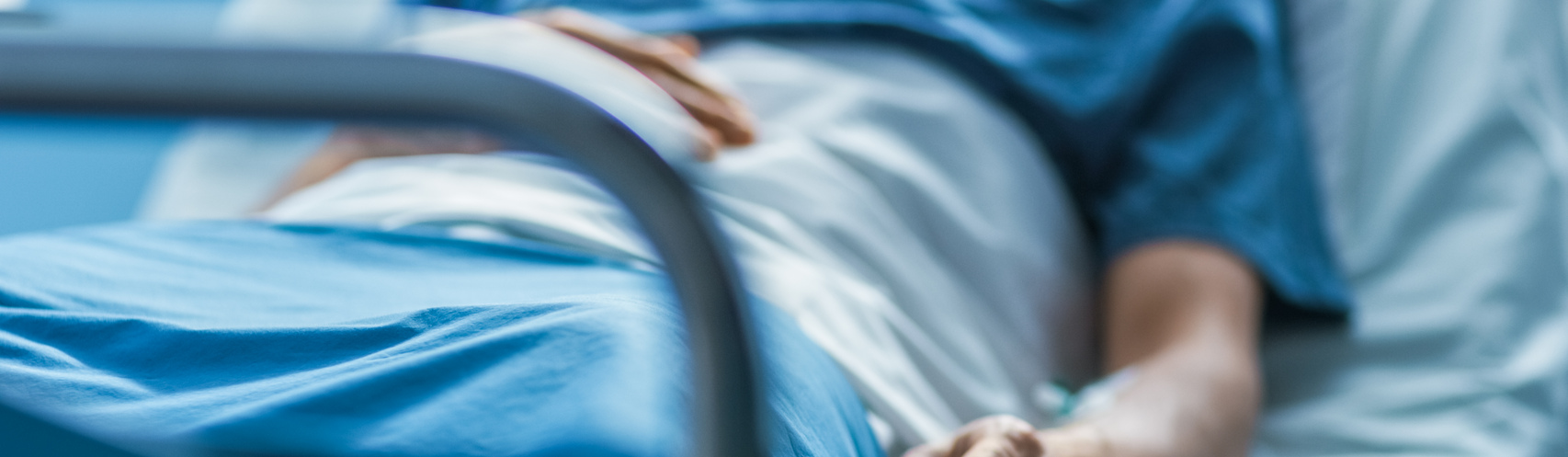 A person is laying in a hospital bed. The picture shows them from the chest down and is angled from the end of the bed. They are wearing a a blood pressure monitor on their finger.