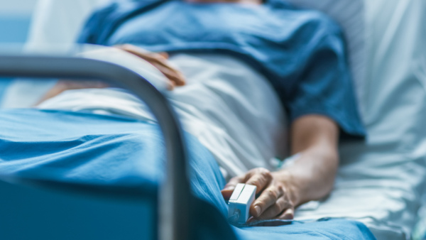 A person is laying in a hospital bed. The picture shows them from the chest down and is angled from the end of the bed. They are wearing a a blood pressure monitor on their finger.